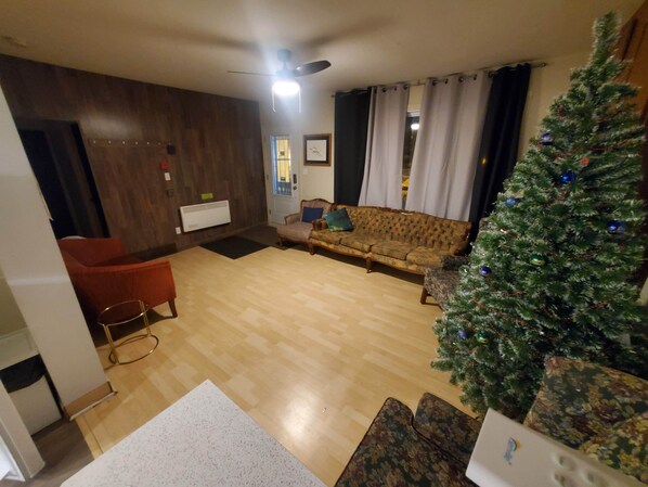 Una sala de estar con piso de madera, un sofá, un sillón rojo, un árbol de Navidad y un ventilador de techo.