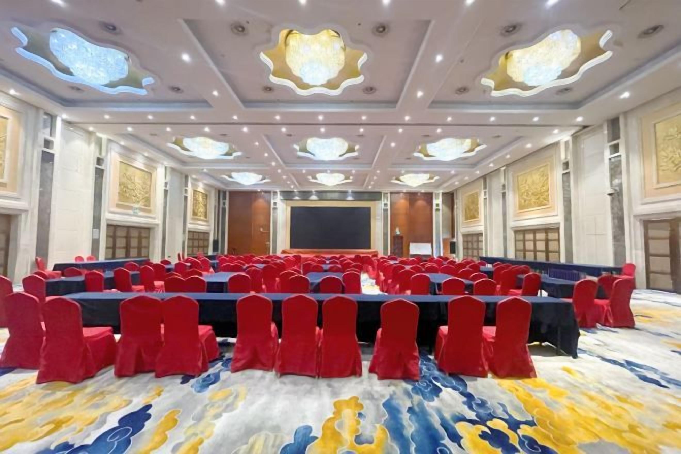 Un salón de conferencias con filas de mesas y sillas, una pantalla grande y detalles ornamentales en el techo.