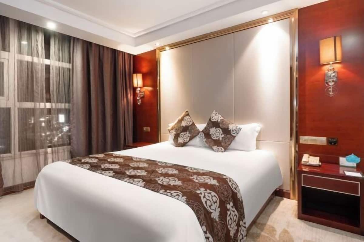 A hotel room with a large bed, a nightstand, a wall-mounted lamp, and a window with curtains.