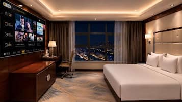 Una habitación de hotel con una televisión grande, una cama, un escritorio, una silla y vistas de la ciudad por la noche.