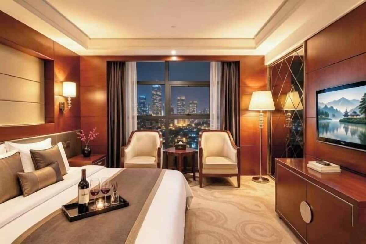 A hotel room with a bed, two armchairs, a TV, a city view, and a tray with a bottle and glasses.