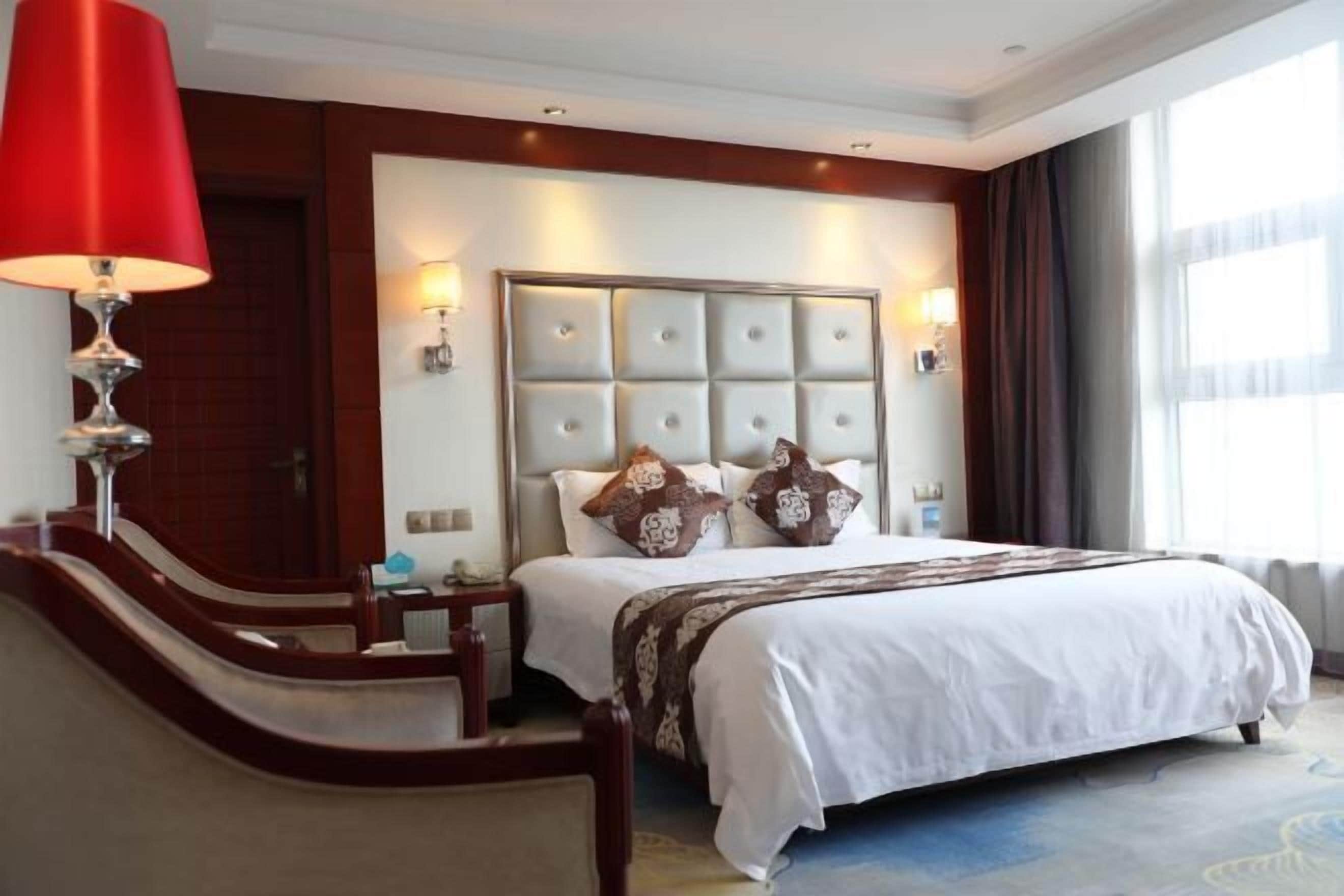 A hotel room with a large bed, a red lamp, a wooden bench, and a view of the outdoors.