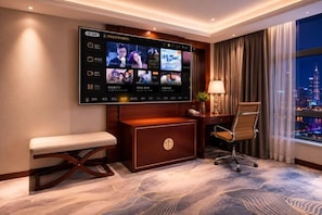 A hotel room with a large flat-screen TV mounted on the wall, a desk with a chair, a bench, and a view of a cityscape through large windows. - Dingsheng International Hotel (Nanchang)