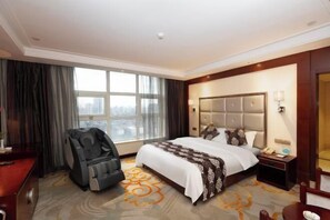 Habitación de hotel con una cama grande, una silla de masaje, una mesita de noche y vistas a la ciudad desde la ventana.