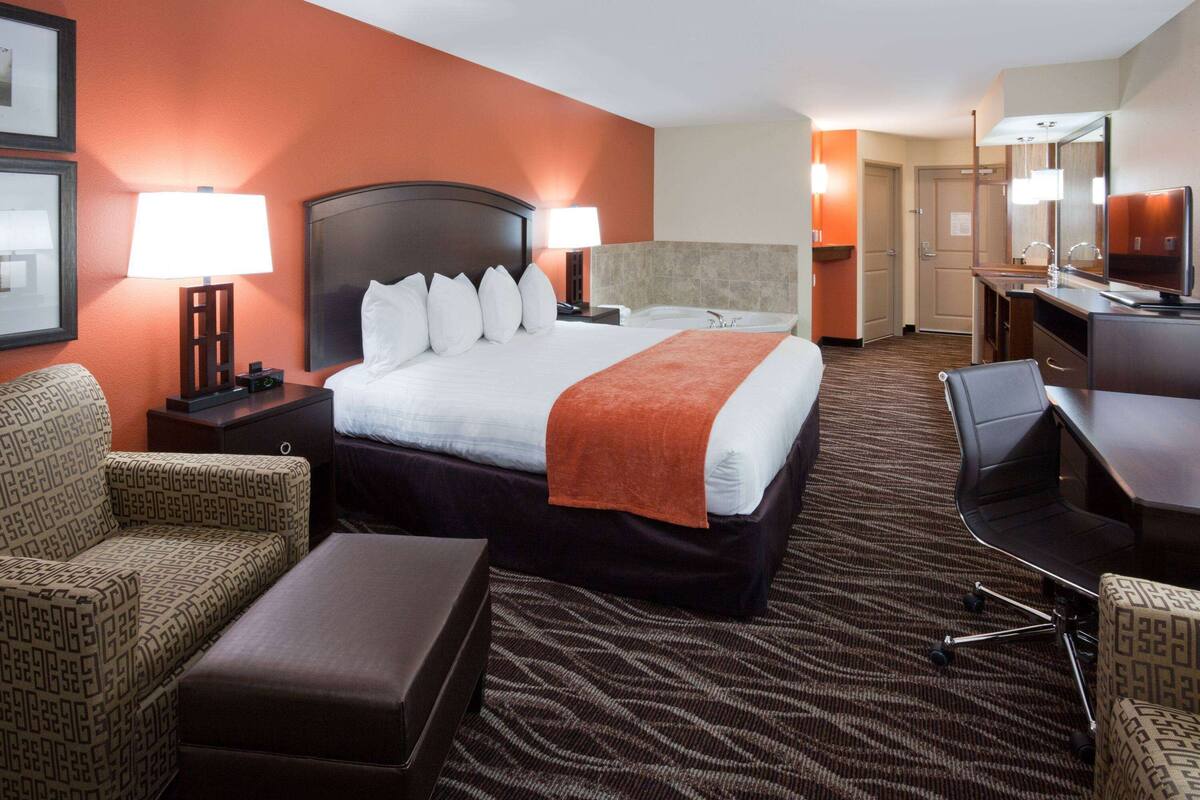A hotel room with a large bed, two bedside lamps, a desk with a chair, a sofa, and a small ottoman.