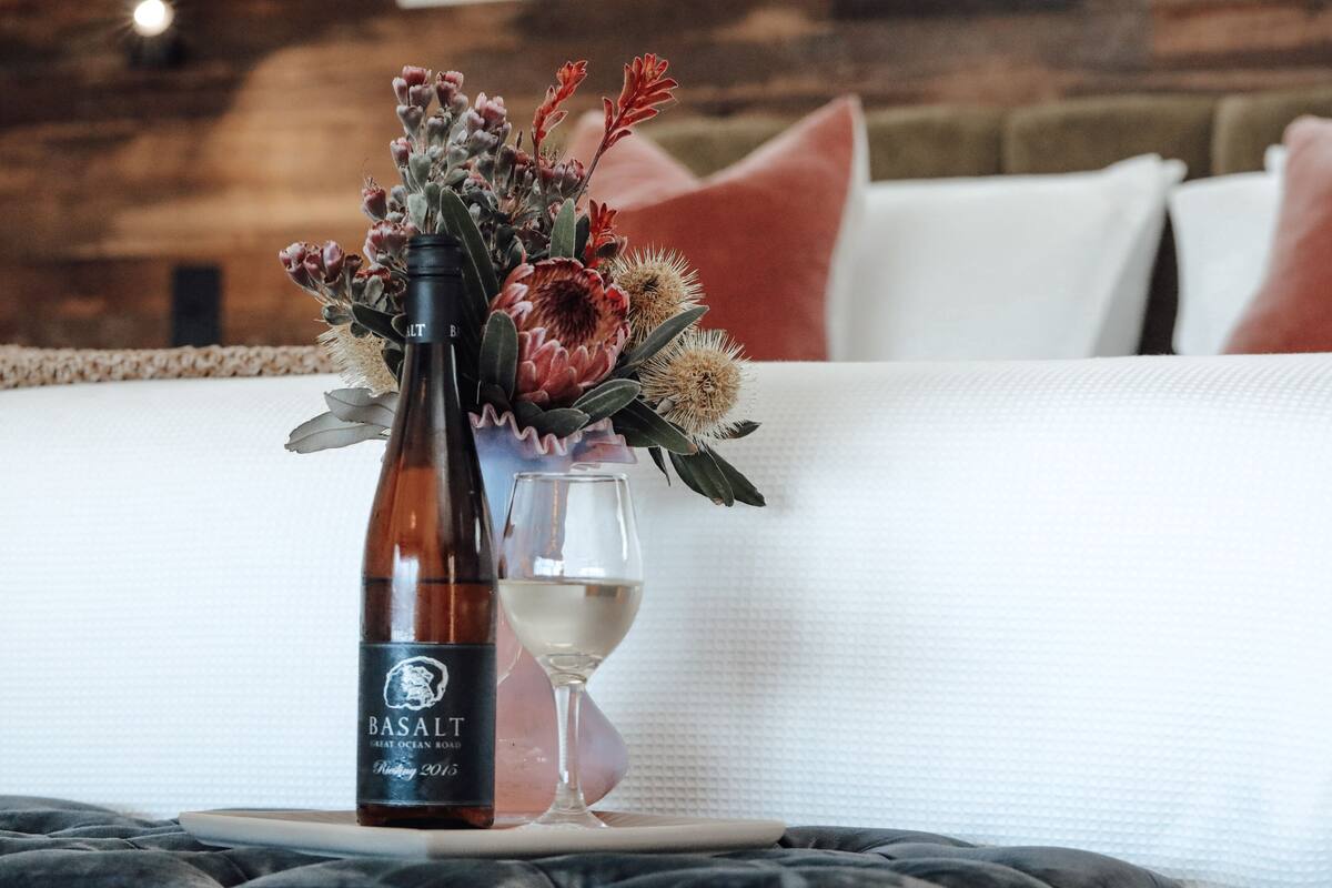 A bottle of Basalt wine, a glass of white wine, and a floral arrangement on a bed with a textured white bedspread.
