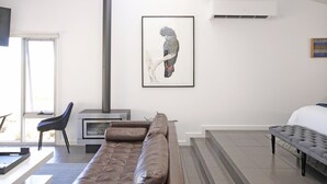 A modern living room with a brown sofa, a fireplace, a framed artwork, and a bed with a white duvet. - Anchors Port Campbell (Port Campbell)