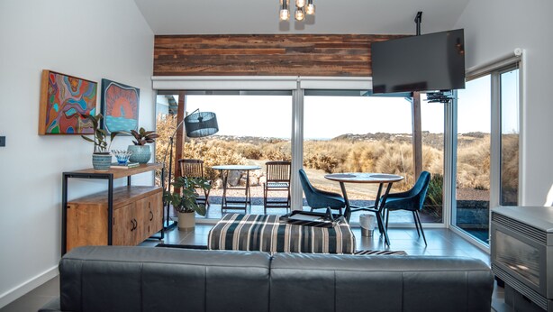 A modern living room with a large sofa, a dining area with chairs and a table, and a television mounted on the wall. - Anchors Port Campbell (Port Campbell)