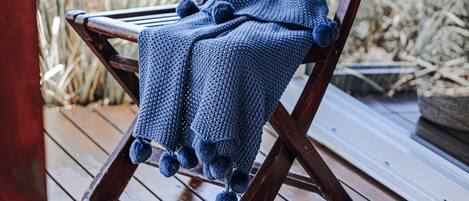 Una silla de madera con una manta azul tejida y pompones, colocada sobre una terraza de madera junto a una ventana con vista a pastos altos.