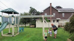 Un parque infantil con tobogán y columpios, una casa al fondo y un césped.