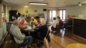 Un grupo de personas sentadas alrededor de una mesa en una habitación con pisos de madera, un ventilador de techo y una ventana que deja entrar luz natural.