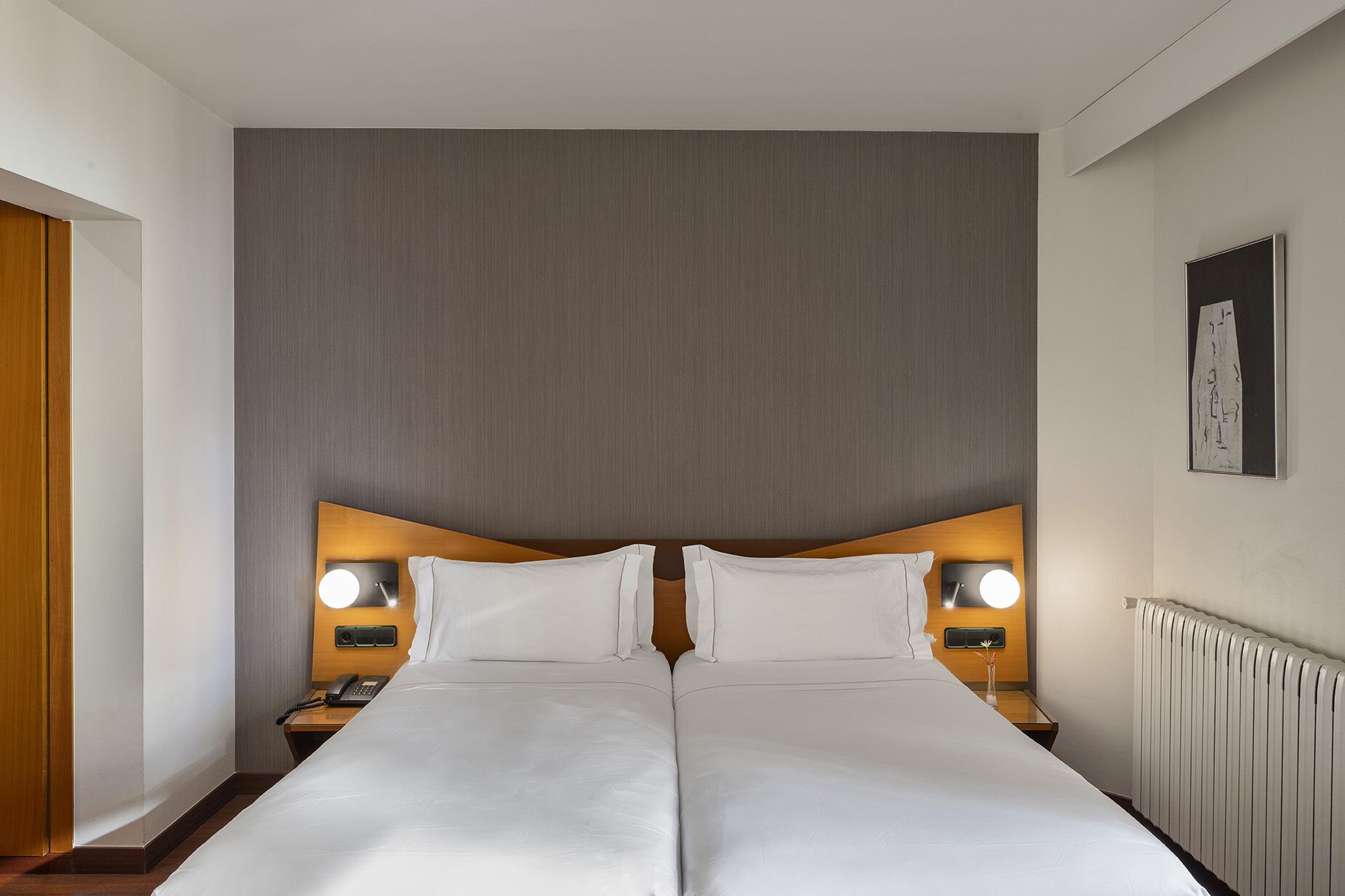 Una cama doble con dos mesitas de noche, cada una con una lámpara, en una habitación de hotel con una pared lisa y un radiador.