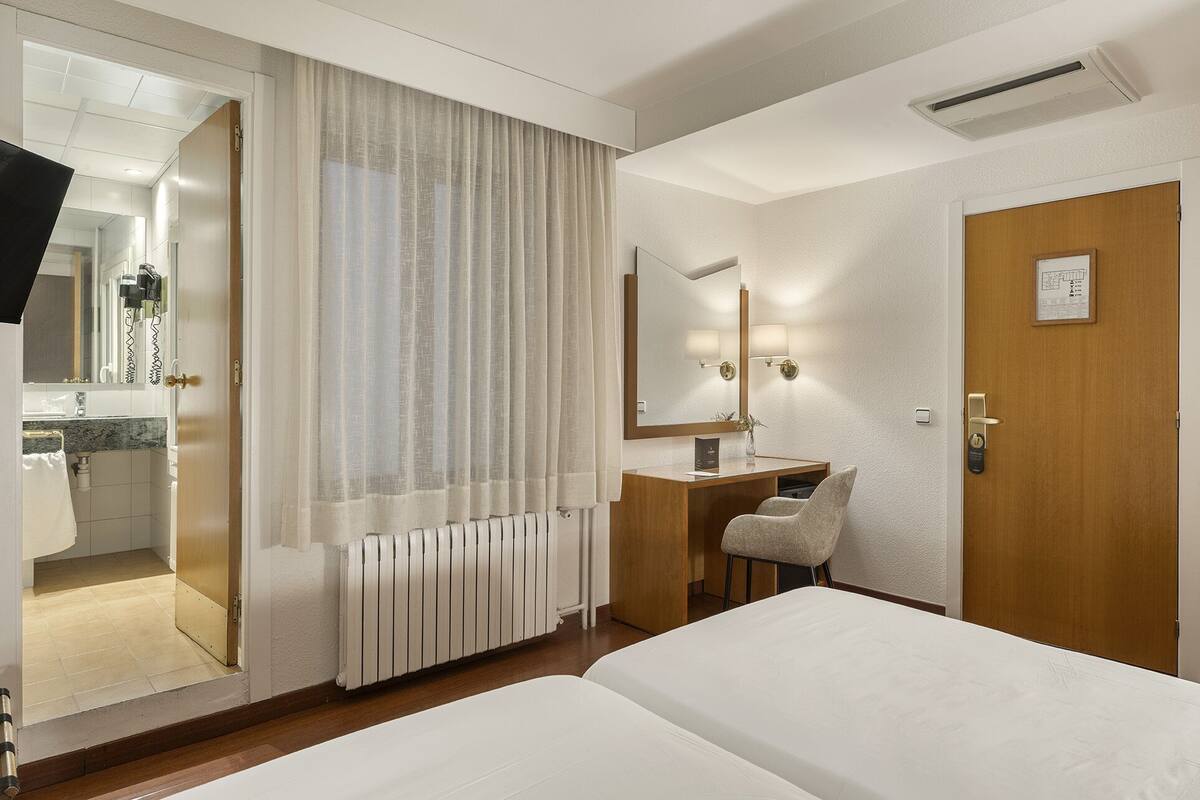 Ein Hotelzimmer mit Bett, Schreibtisch mit Stuhl, Spiegel und einem durchgegangenen Tür sichtbaren Badezimmer.