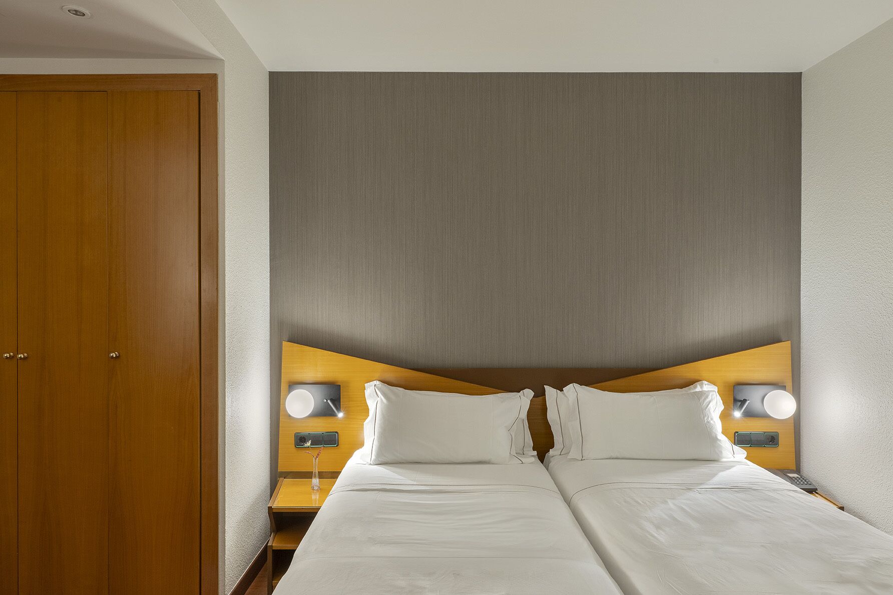 Habitación de hotel con cama doble, dos mesitas de noche con lámparas, cabecera de madera y una puerta.