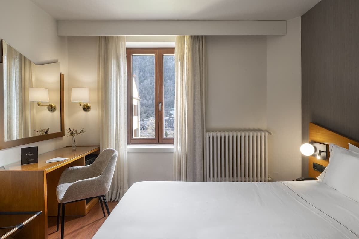 Ein Hotelzimmer mit Bett, Schreibtisch mit Stuhl, großem Spiegel, Fenster mit Vorhängen und Heizkörper.