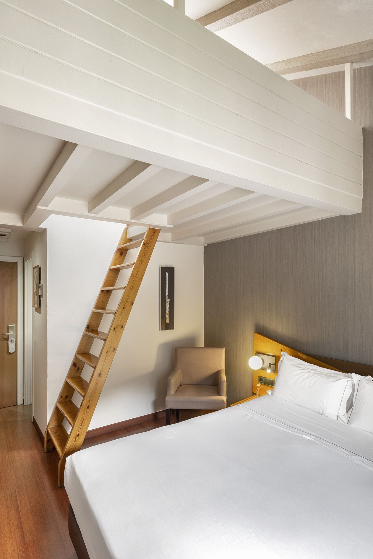 A hotel room with a wooden ladder, a bed, a chair, and a nightstand with a lamp.