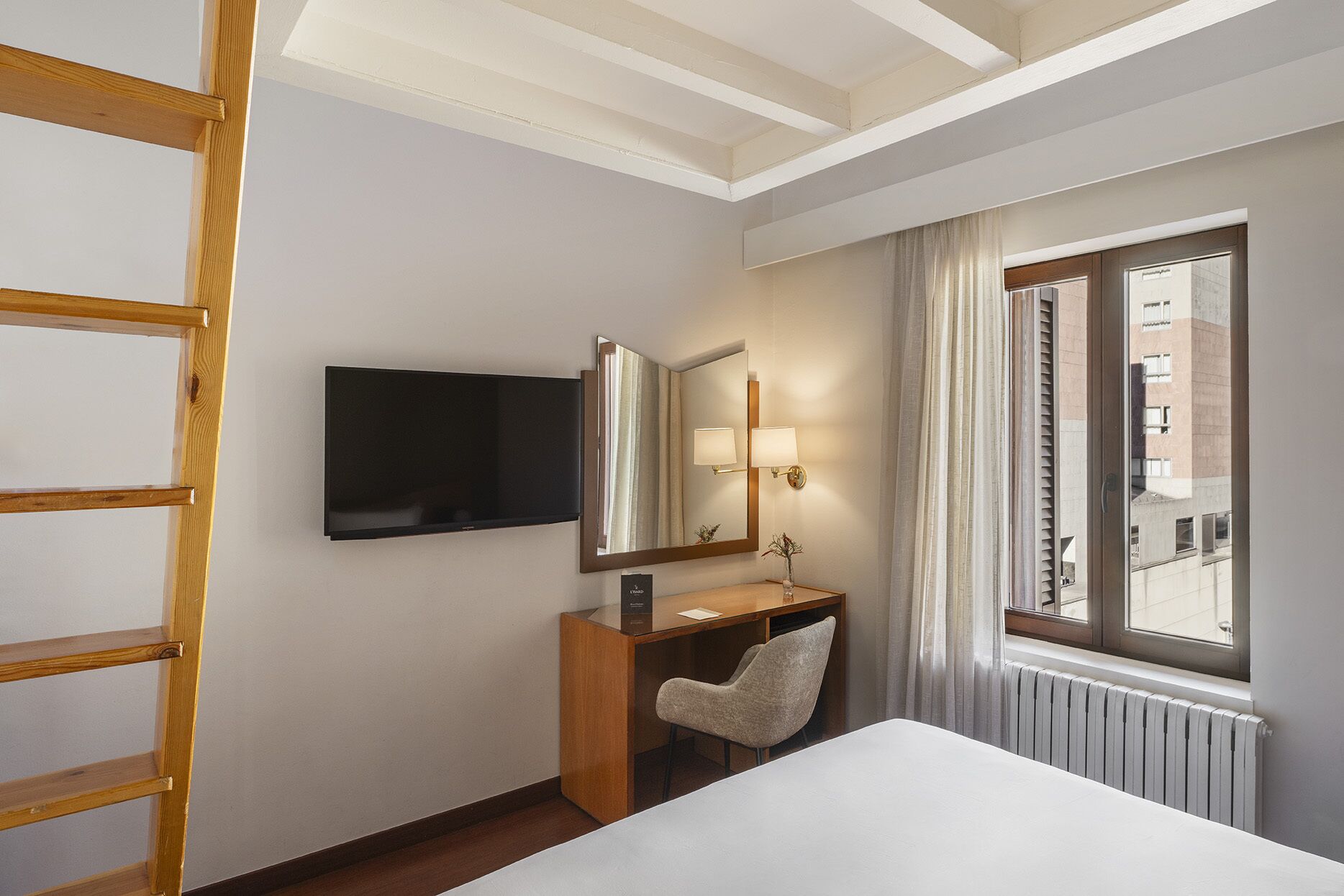 Una habitación de hotel con cama, escritorio y silla, televisor montado en la pared, ventana con cortinas y radiador.