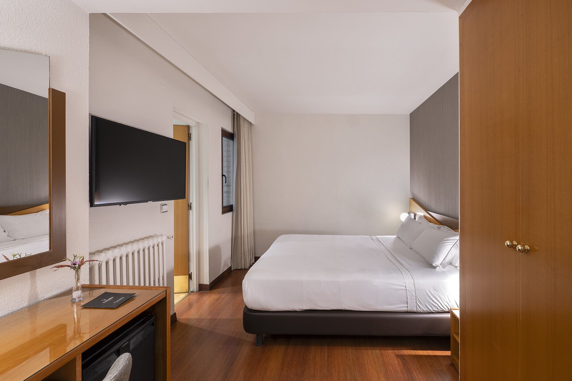 Habitación de hotel con cama, televisor en la pared, escritorio y silla, y un radiador.