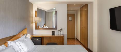 Habitación de hotel con una cama grande, un escritorio con lámpara, un espejo y un televisor montado en la pared.