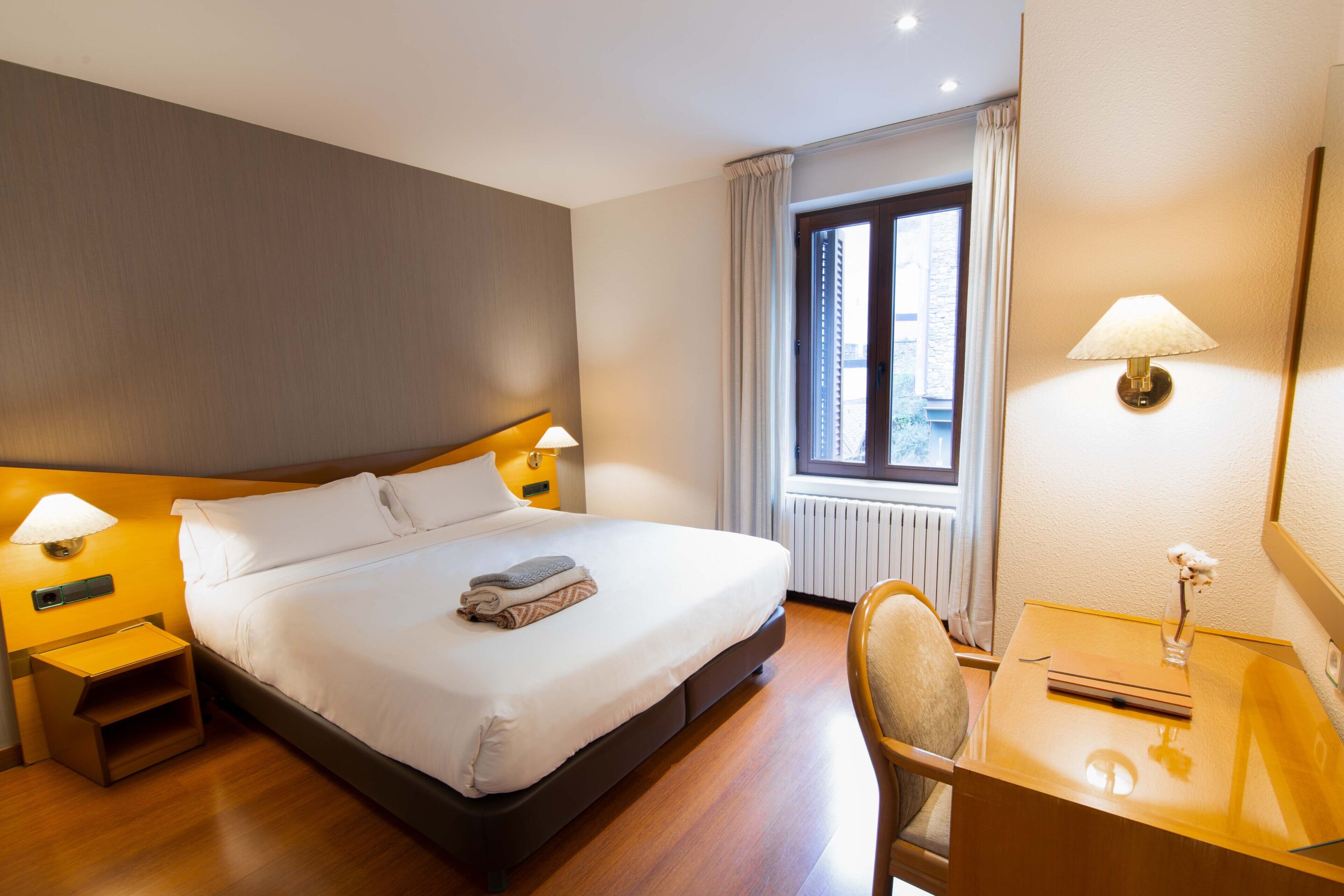 Habitación de hotel con cama, lámparas de noche, escritorio con lámpara, una silla, ventana con cortinas y un radiador.