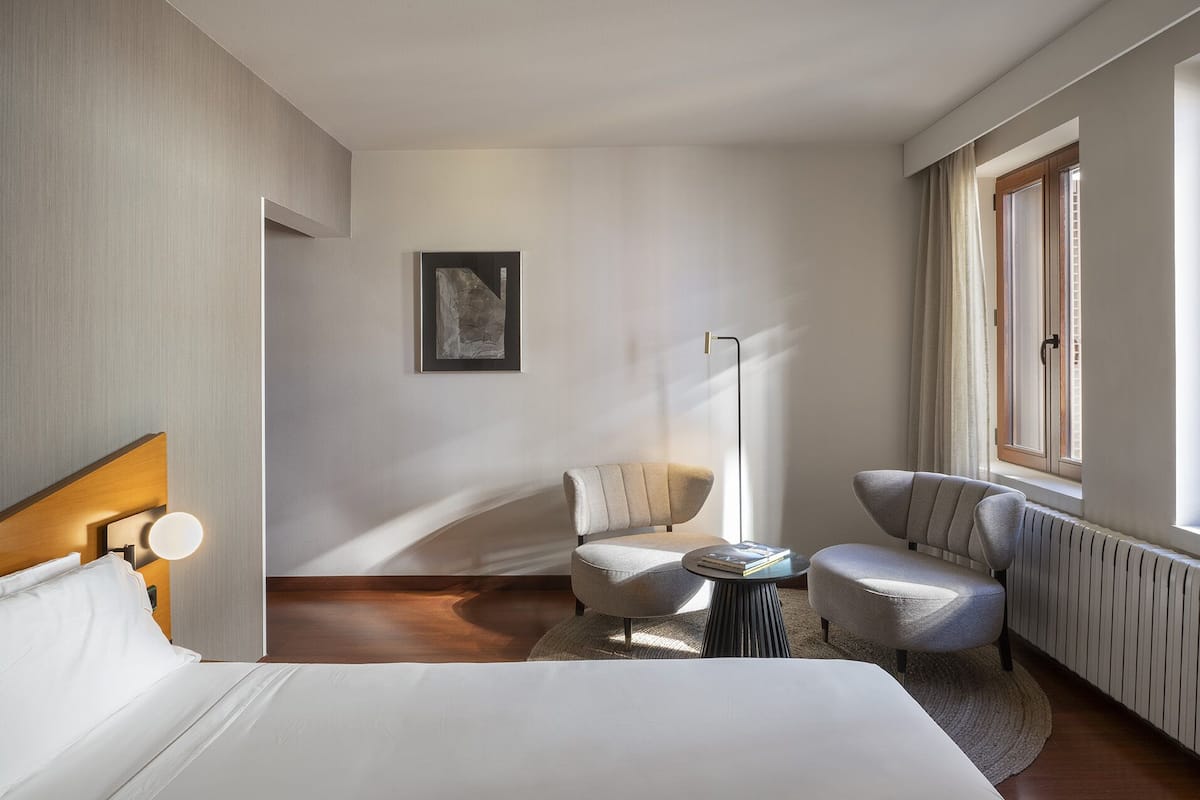Ein modernes Hotelzimmer mit einem Bett, zwei Sesseln, einem kleinen runden Tisch, einer Stehlampe und einem Bild an der Wand.