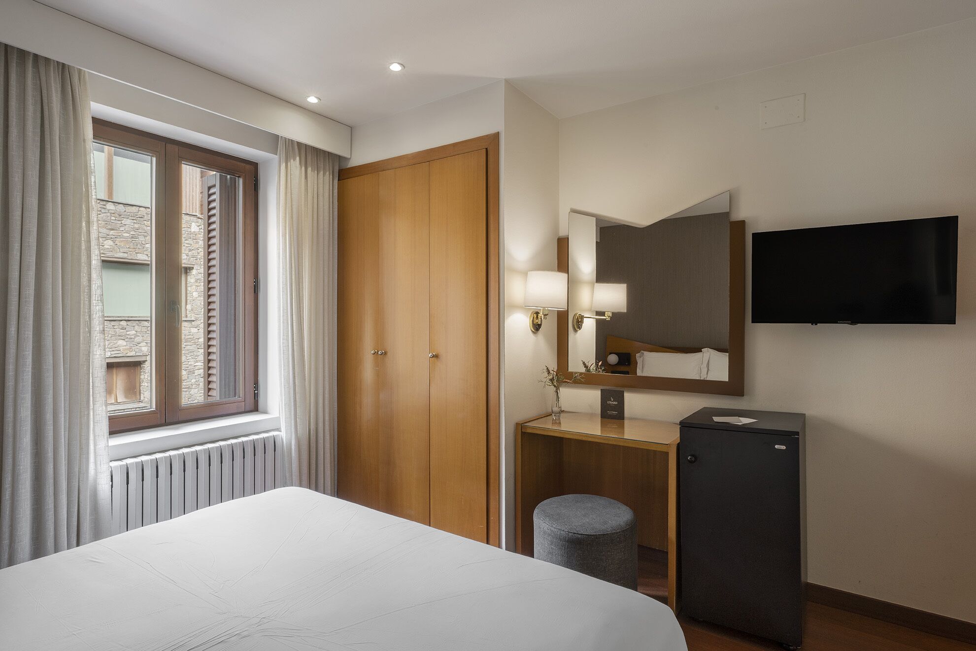 A hotel room with a bed, a wooden wardrobe, a desk with a mirror, a television, and a radiator.