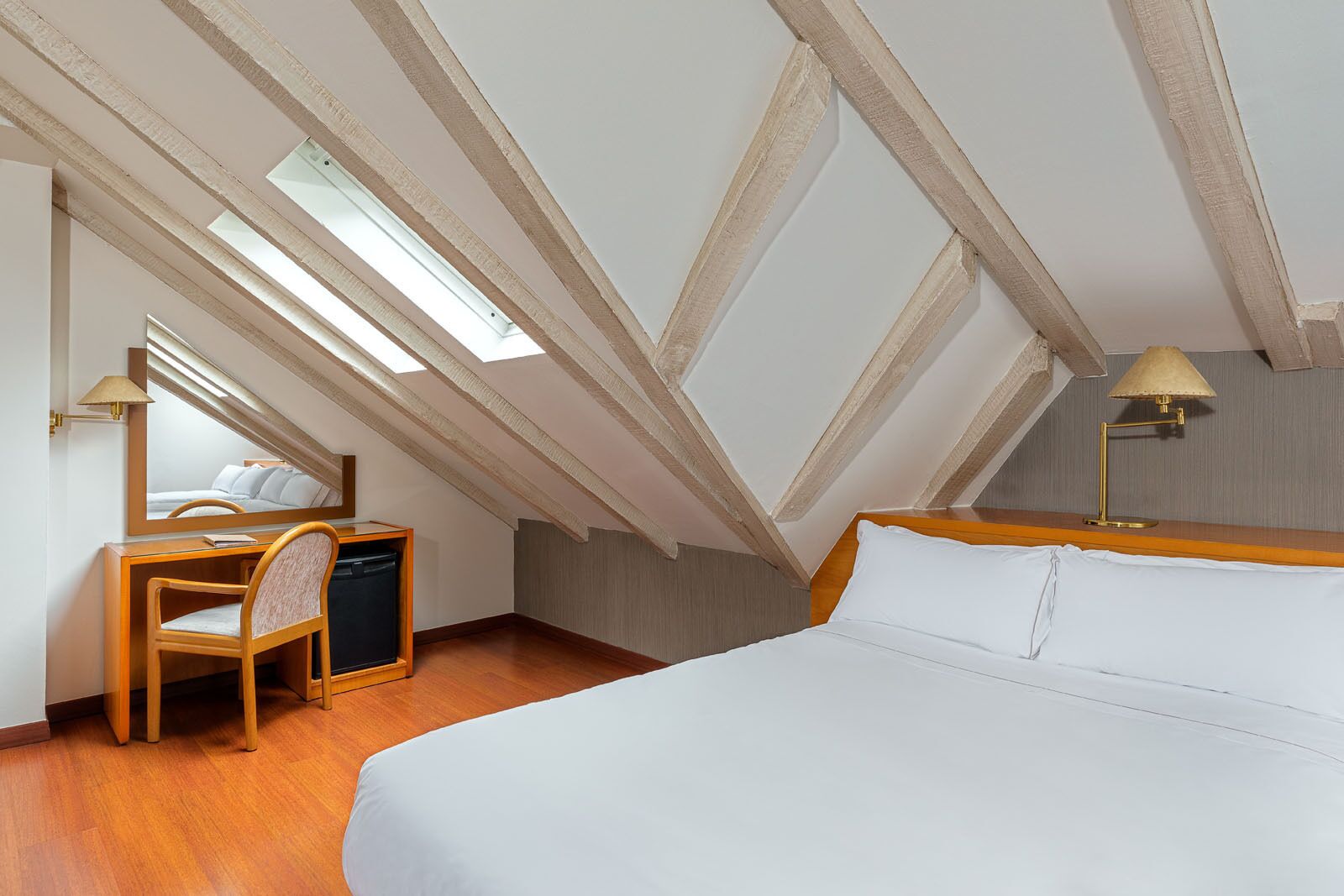 A bedroom with a sloped ceiling, a bed, a desk with a chair, and a mirror.