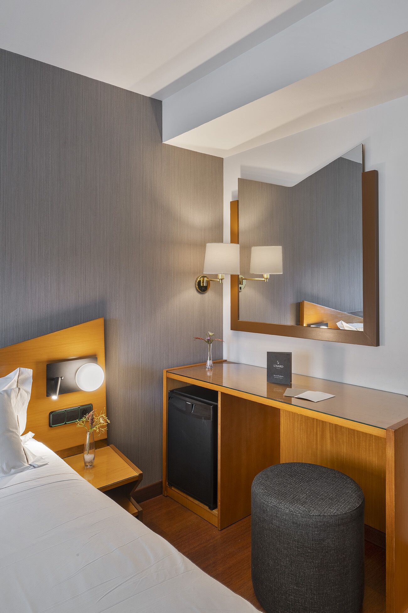 A hotel room with a bed, bedside table, lamp, desk, mirror, and a small ottoman.