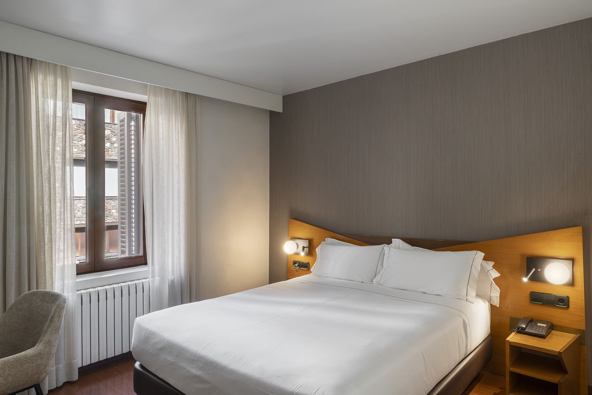 A hotel room with a large bed, a chair, a bedside table with a lamp, and a window with curtains.