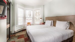 Quarto de hotel com duas camas, uma mesa de cabeceira vermelha, um abajur e uma janela com persianas.