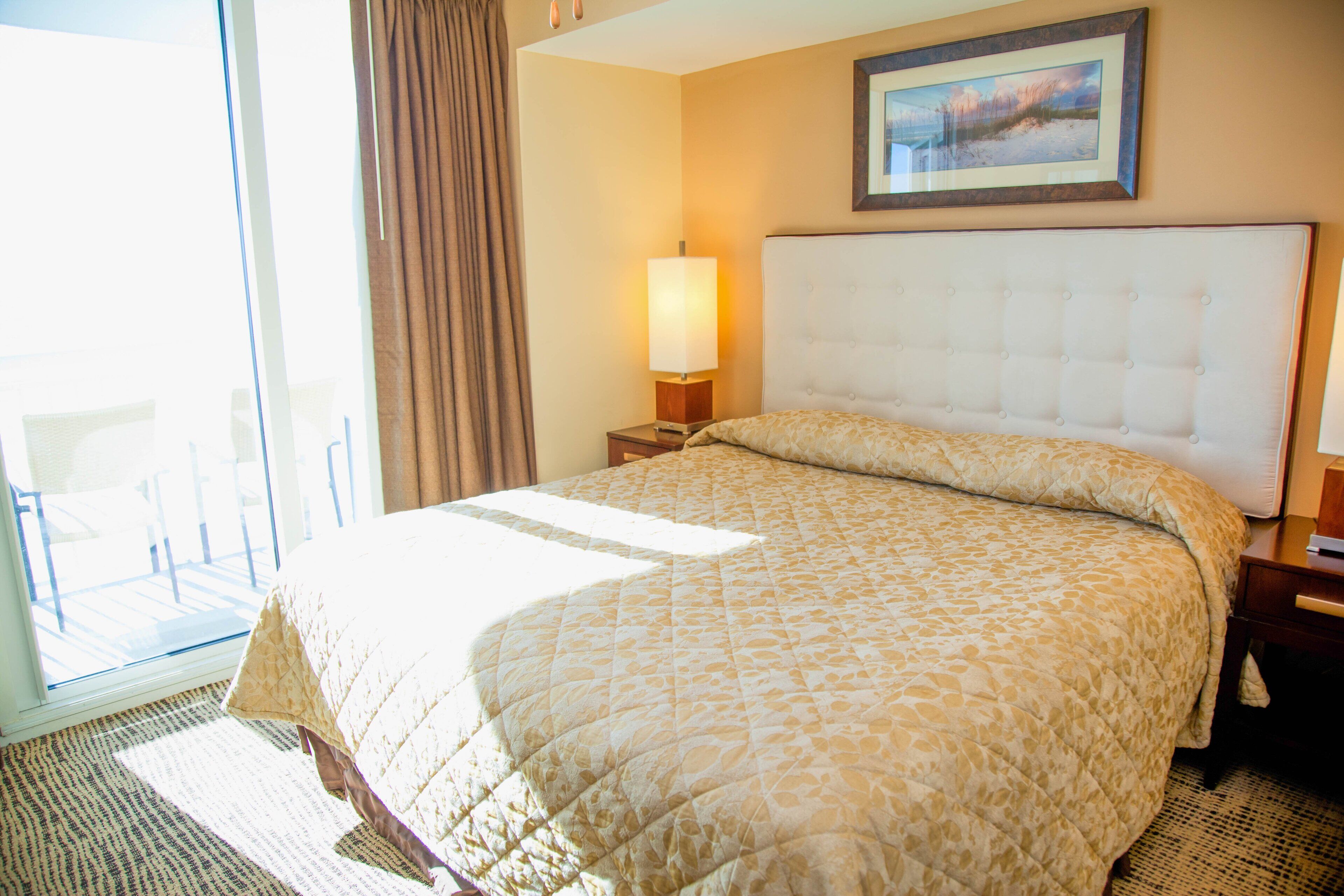 A hotel room with a large bed, a nightstand with a lamp, a window with curtains, and a framed picture on the wall.