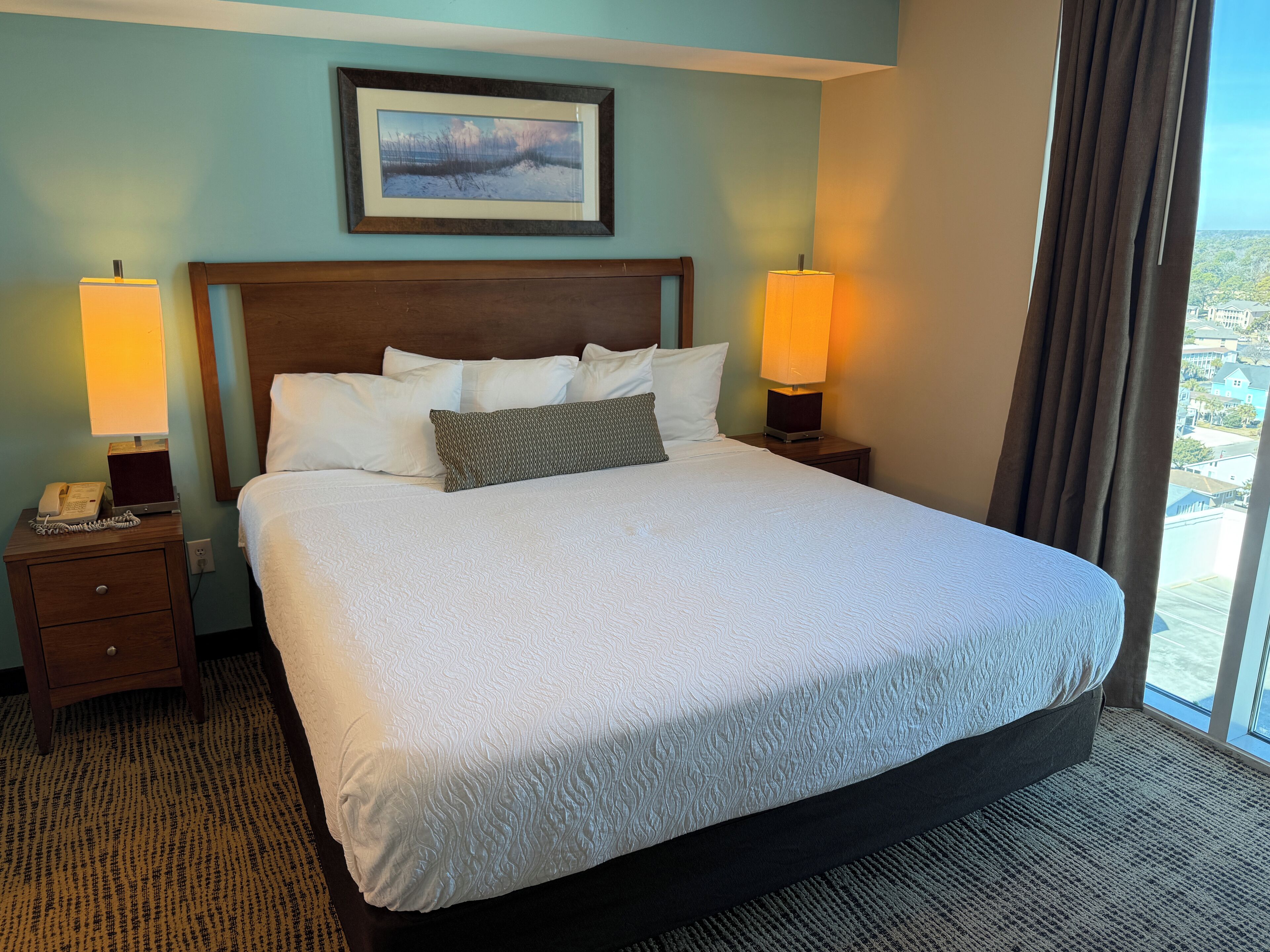 A hotel room with a large bed, two bedside lamps, a framed picture on the wall, and a view of the city through the window.