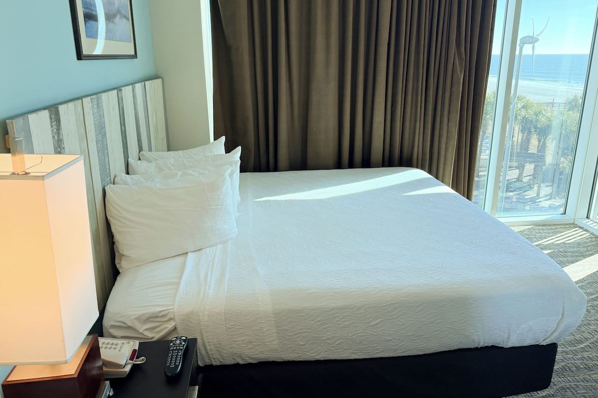 A hotel room with a large bed, a bedside lamp, a remote control, and a view of the ocean through a large window.