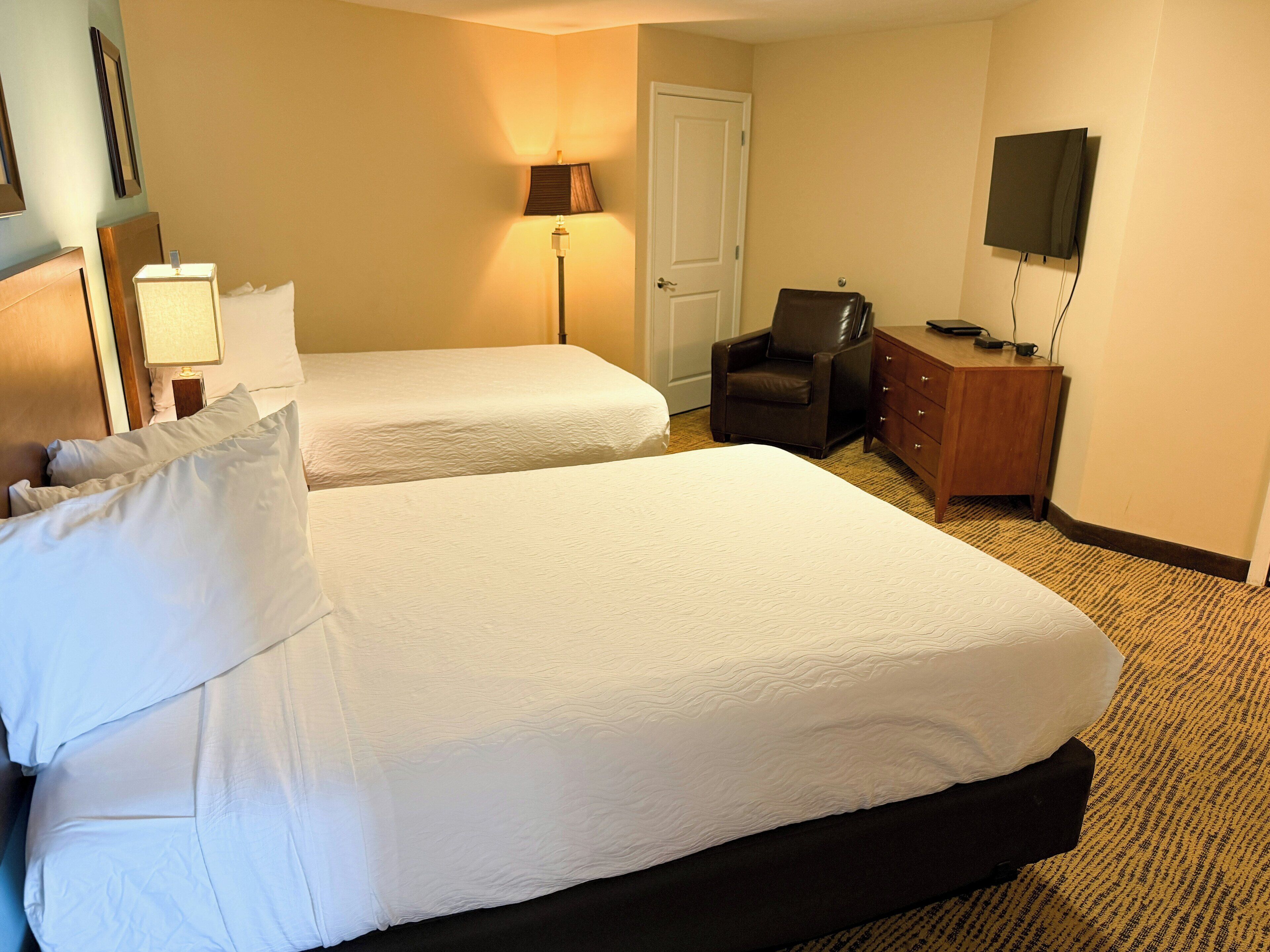 A hotel room with two beds, a desk, a chair, a lamp, and a television.