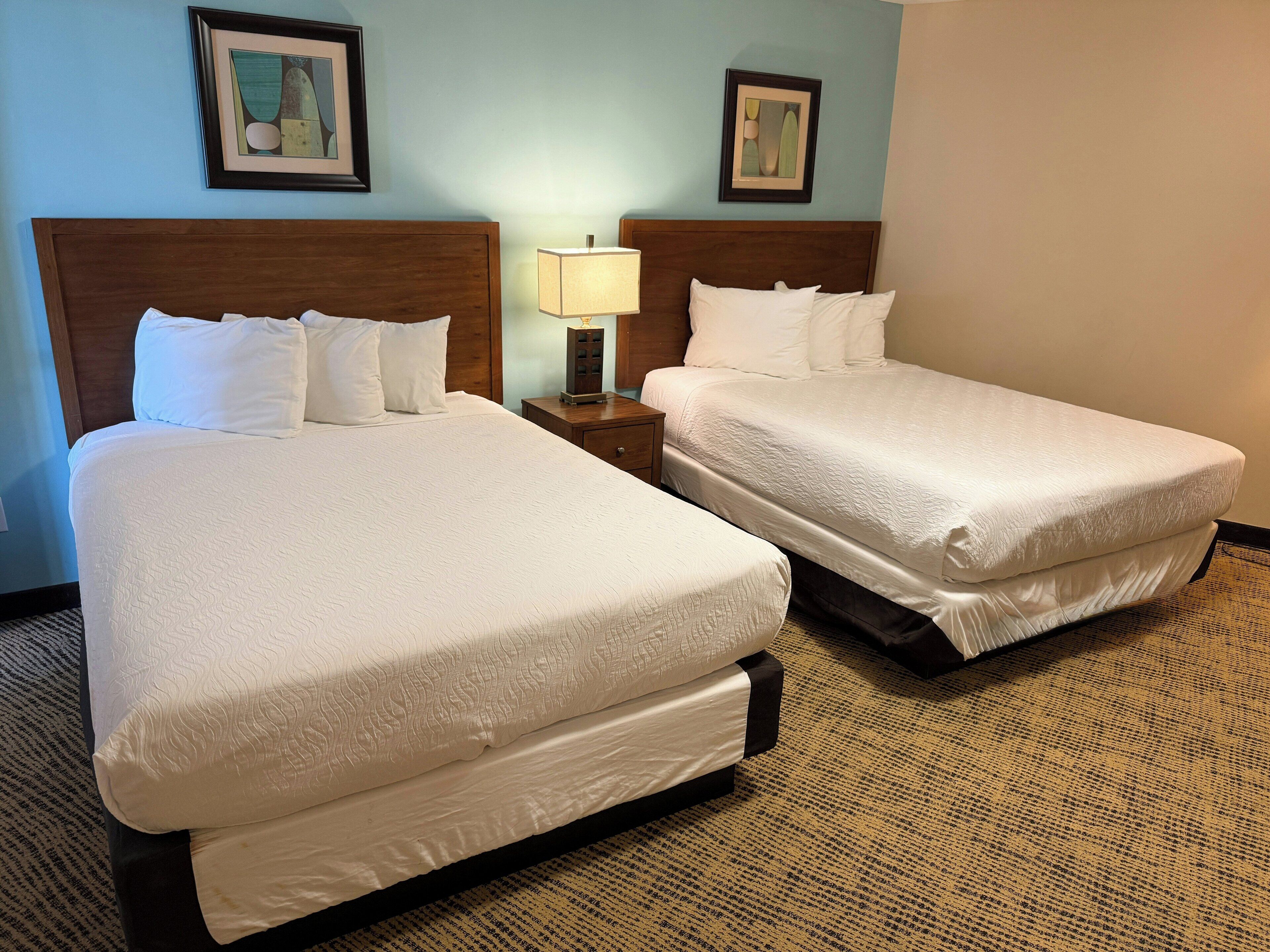 Two double beds with white linens, a nightstand with a lamp, and two framed artworks on the wall.