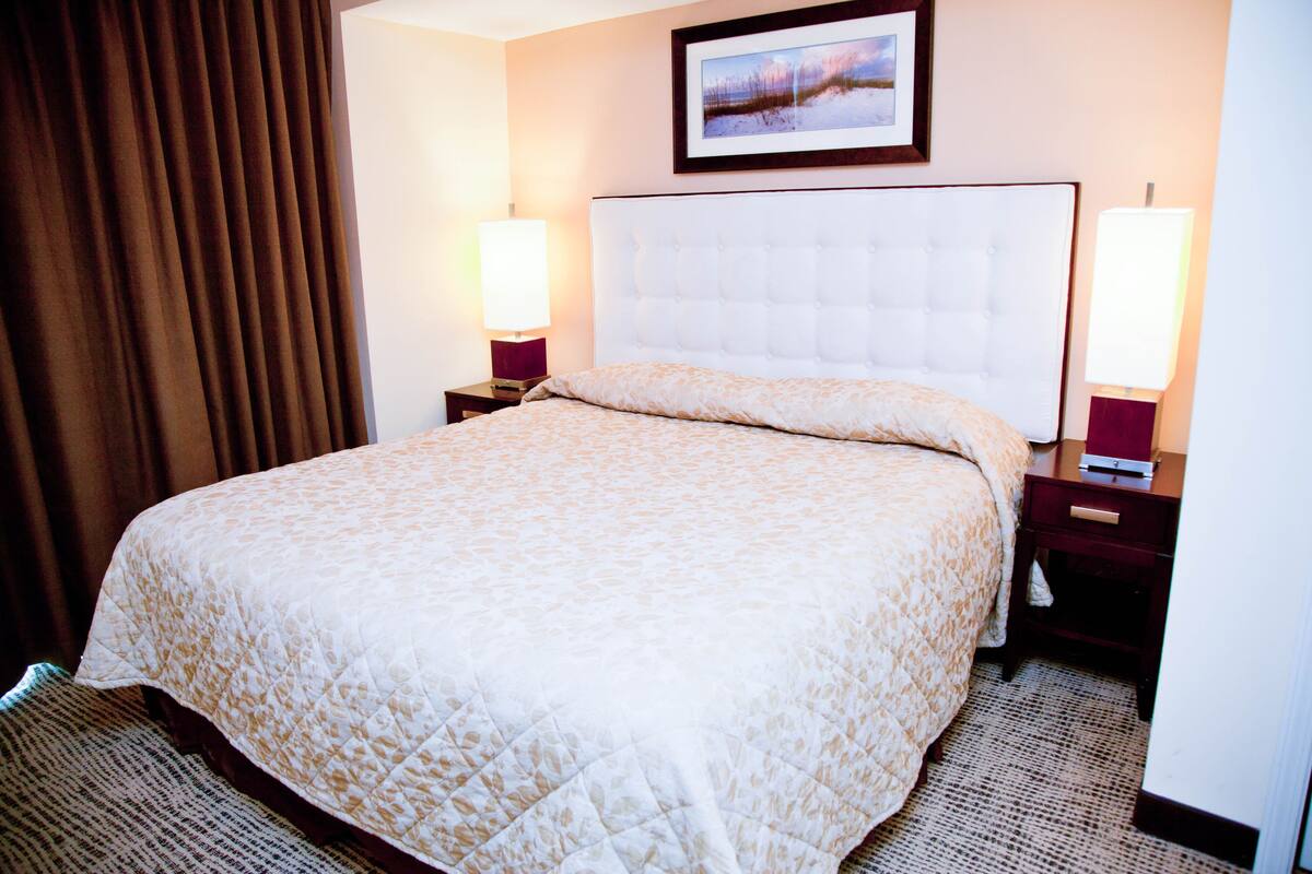 A hotel room with a large bed, two bedside tables with lamps, a framed picture on the wall, and a window with curtains.