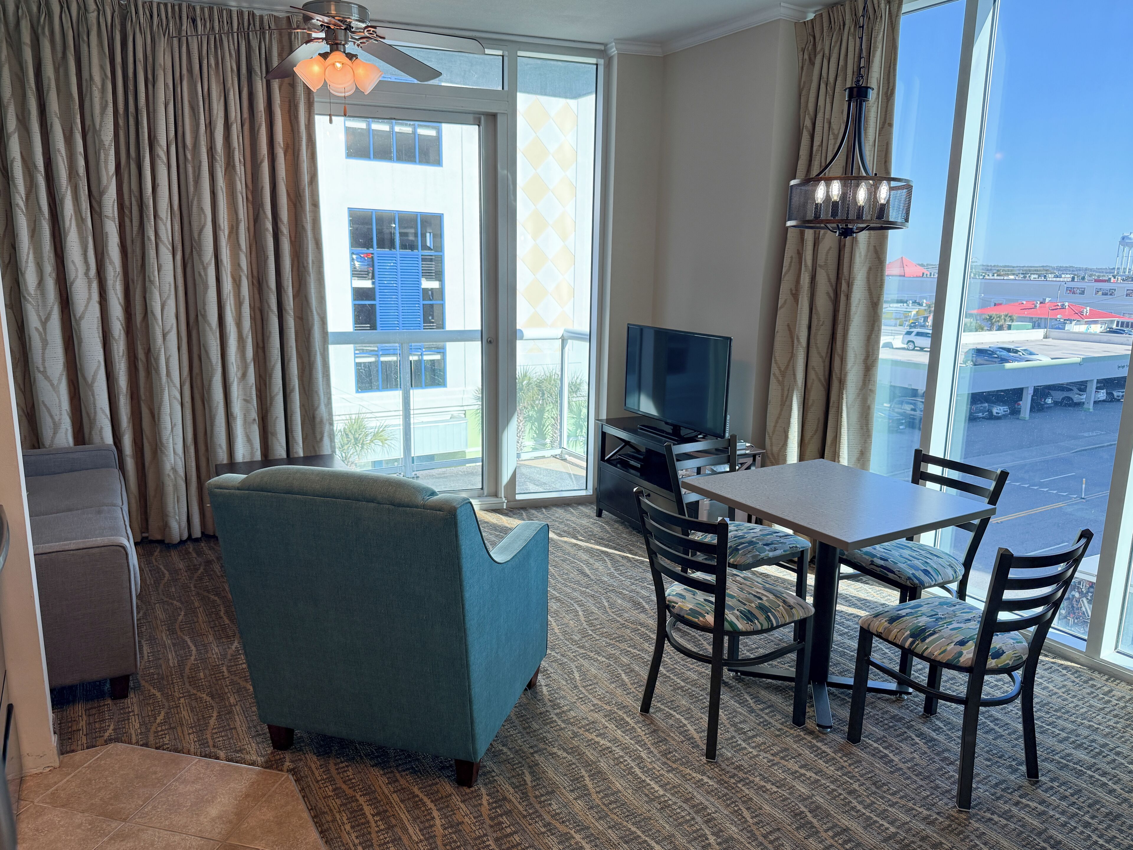 A hotel room with a sofa, a dining table with chairs, a television, and a view of the city through large windows.