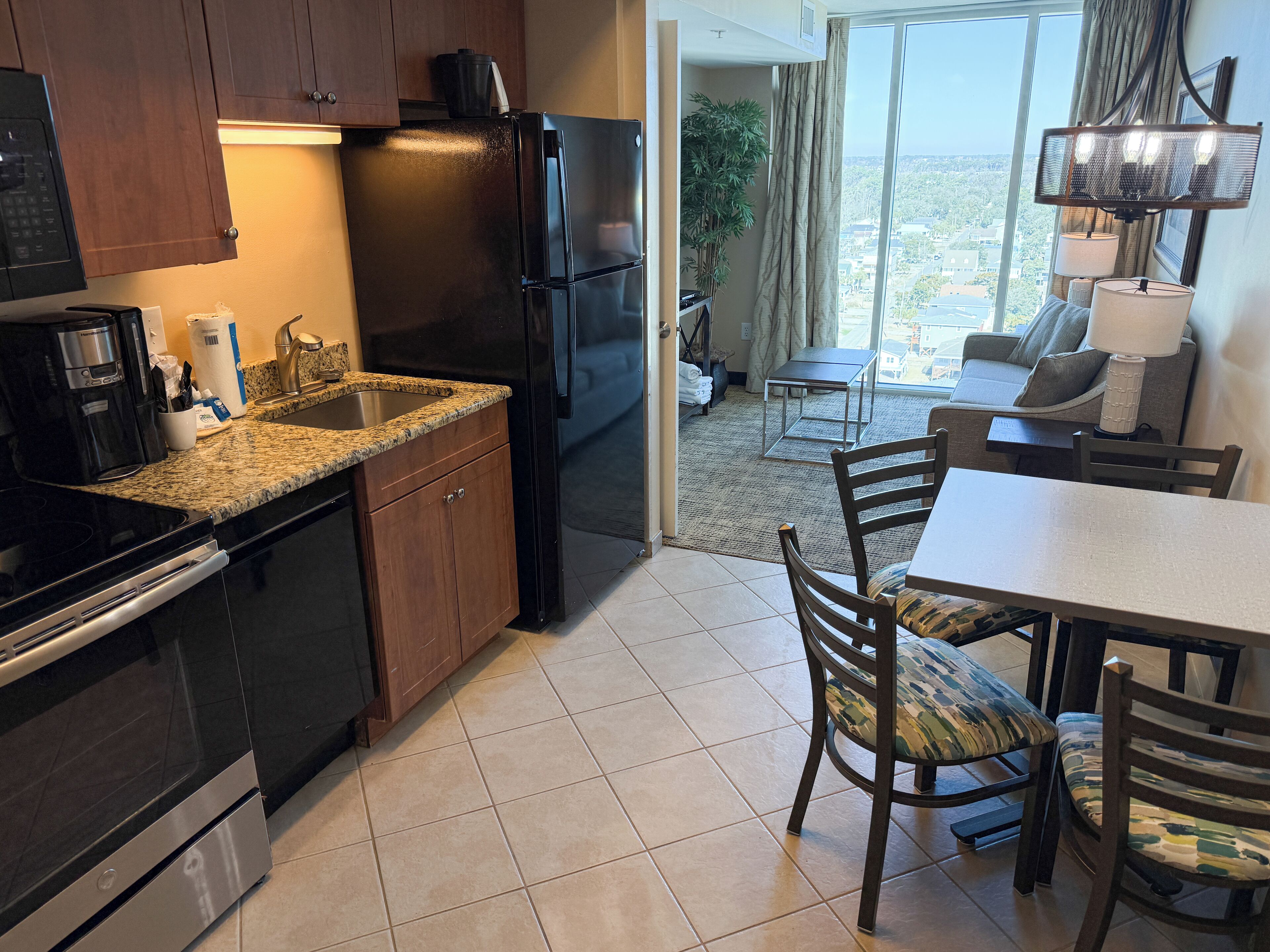 A modern kitchen with granite countertops, a black refrigerator, and wooden cabinets. There is a dining area with a table and chairs, and a view of the city through large windows.