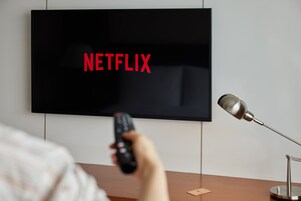 Un televisor con la palabra "NETFLIX" en la pantalla, un control remoto en la mano y una lámpara de escritorio a un lado.