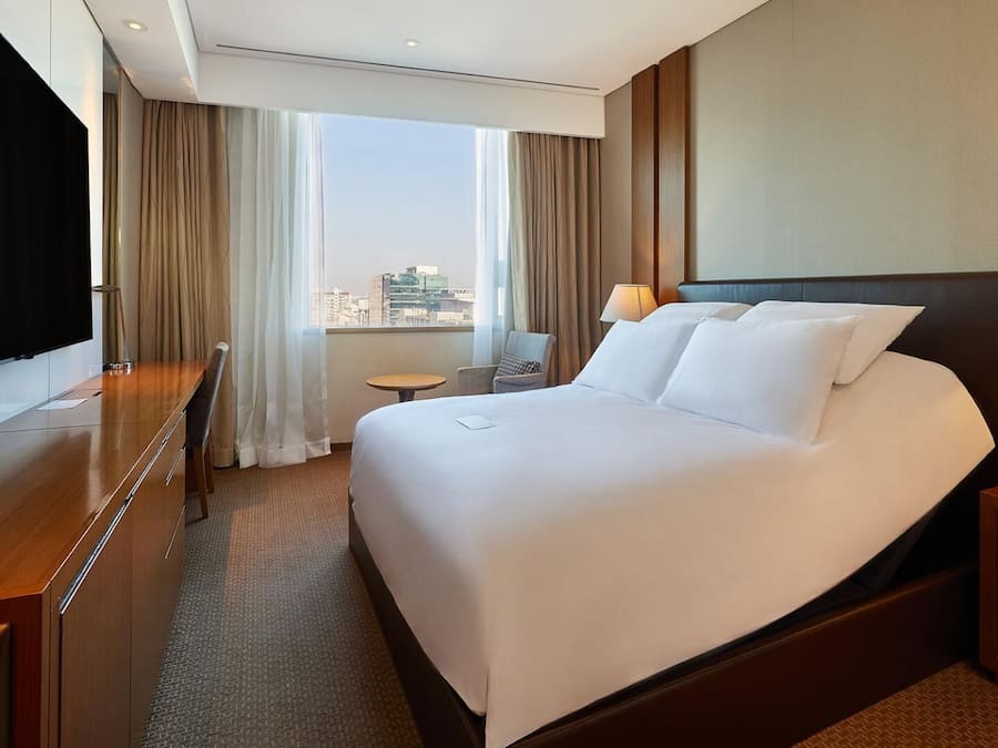 Habitación de hotel con una cama grande, un escritorio, una silla, un televisor y vistas a la ciudad.