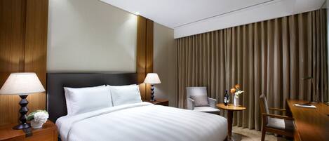 A hotel room with a large bed, two bedside tables with lamps, a small round table with a chair, and a large window with curtains.