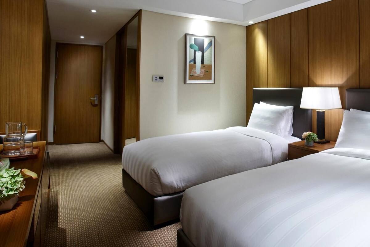 Habitación de hotel con dos camas, una mesita de noche con lámpara, un cuadro en la pared y una puerta que da a otra habitación.