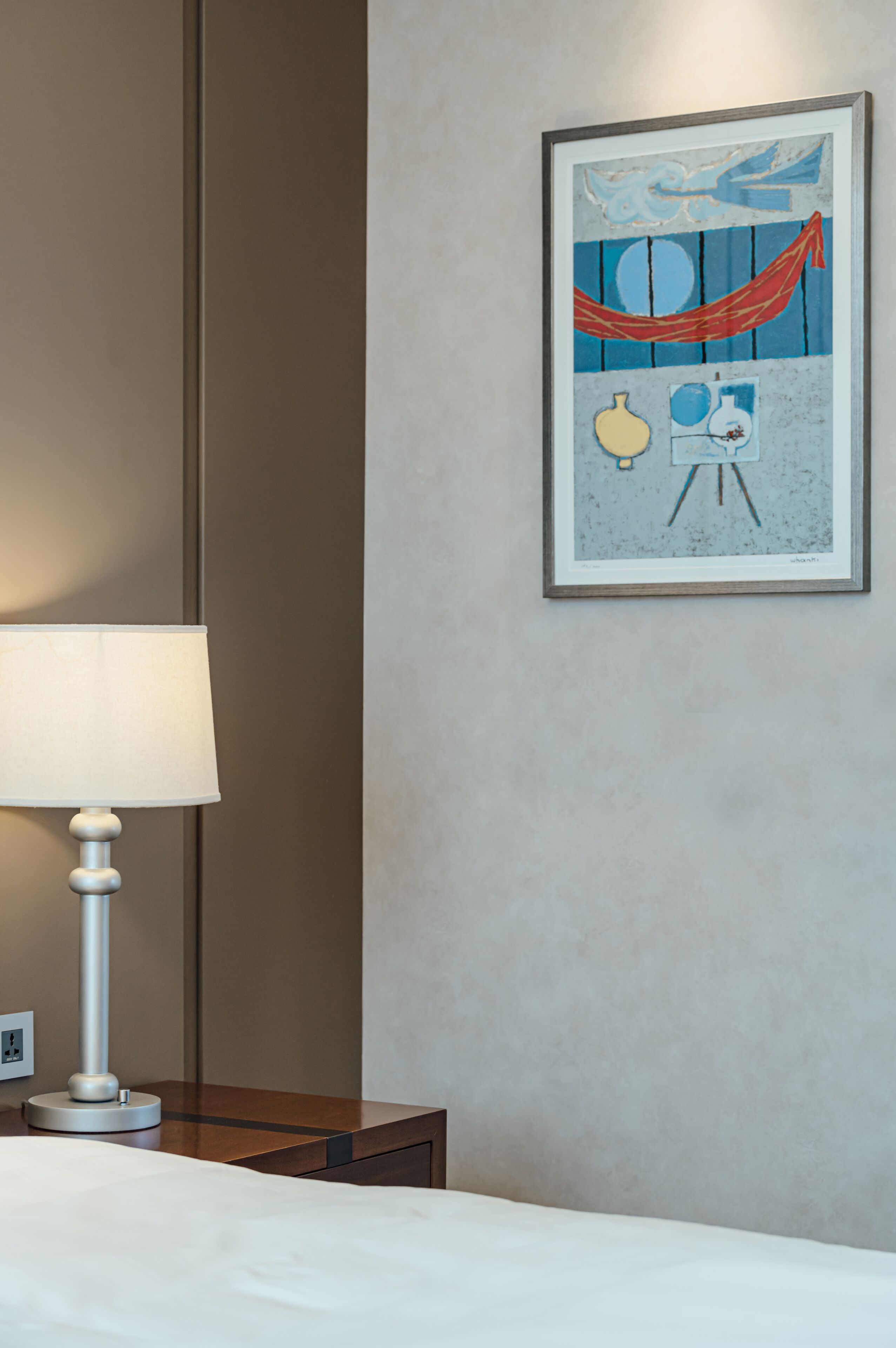 A hotel room with a bed, a bedside table, a lamp, and a framed artwork on the wall.