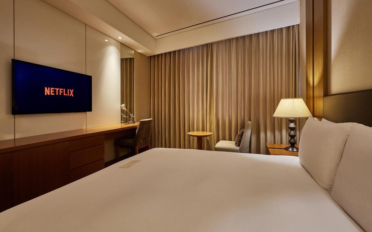 Una habitación de hotel con cama, un televisor mostrando 'NETFLIX', un escritorio, una silla, una lámpara y una cortina.