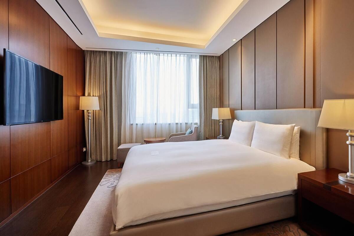 Una habitación de hotel moderna con una cama grande, dos lámparas de noche, un televisor de pantalla plana y un ventanal con cortinas.
