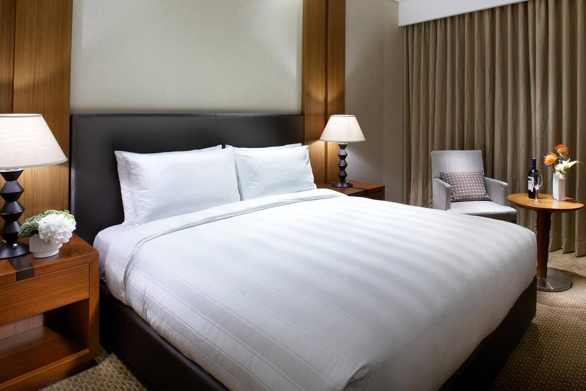 Una habitación de hotel con una cama grande, dos mesitas de noche con lámparas, una silla, una pequeña mesa con un florero y botellas, y un telón de fondo de cortina.