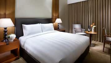 A hotel room with a large bed, two bedside tables with lamps, a chair, a small table with a vase and bottles, and a curtain backdrop.