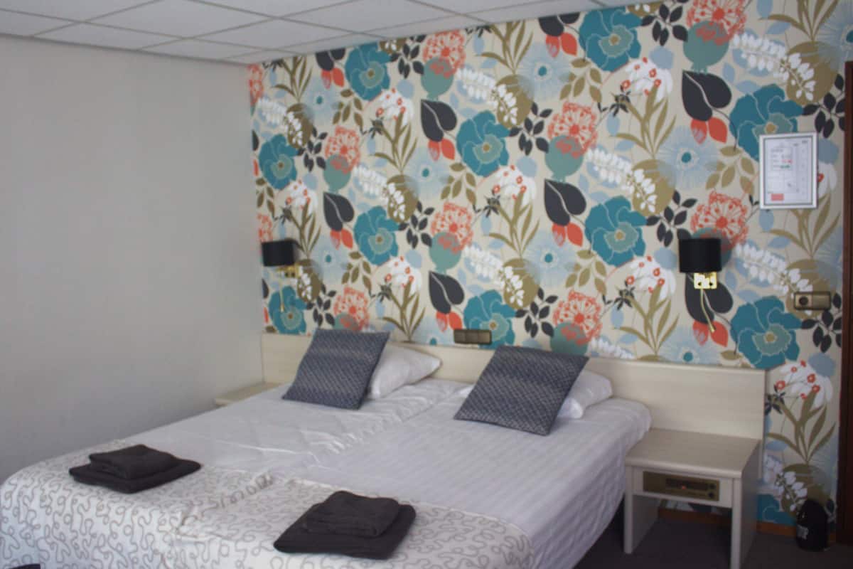 A hotel room with a floral wallpaper, a double bed with two pillows, a bedside table with a lamp, and a small nightstand with a lamp.