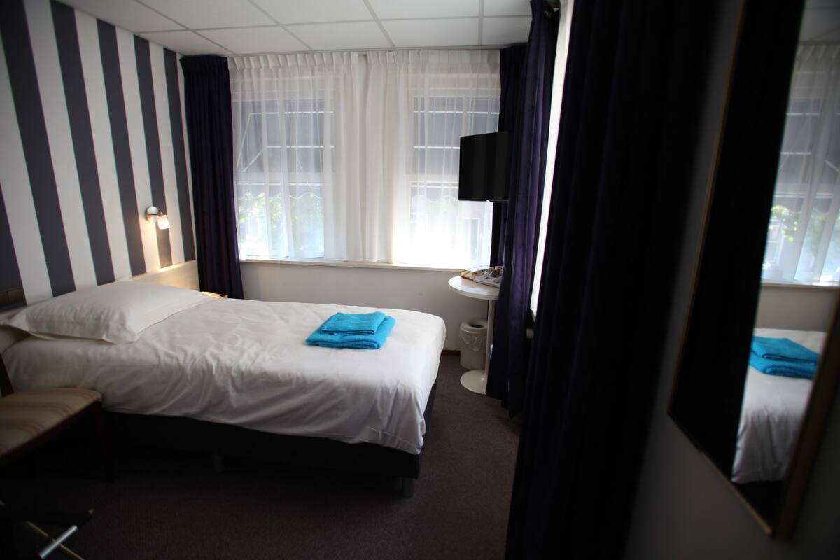A hotel room with a bed, a bedside table, a mirror, a window with curtains, and a wall-mounted TV.