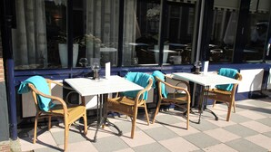 Een terras met rieten stoelen en witte tafels, met een geruit vloertje en grote ramen voorzien van gordijnen.